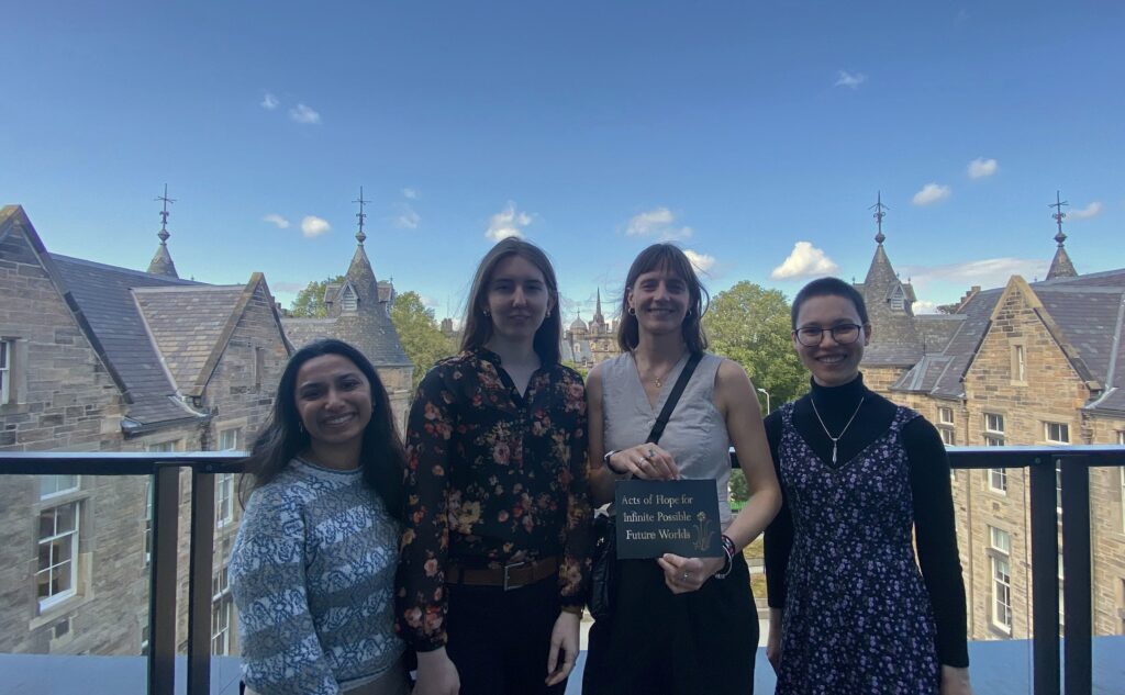 Image of four people standing in a line smiling at the camera. One is holding a piece of card that reads 'Acts of Hope for Infinite Possible Futures Worlds'